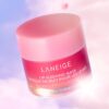 Alt text: A pink jar of LANEIGE Lip Sleeping Mask sits on a white surface, surrounded by fresh berries. The jar is open, revealing the creamy texture of the mask. The product promises to rejuvenate lips overnight, providing deep hydration and leaving them soft and supple.