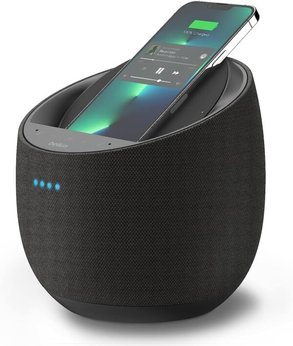 A compact, circular smart speaker in a sleek, modern design, with a built-in wireless charging pad on top. The speaker's exterior features a smooth black finish with a subtle LED indicator light, indicating its operational status. The base of the speaker includes a mesh covering, allowing sound to project outwards. The wireless charging pad has a slight indentation to securely hold a smartphone or other Qi-compatible devices. The overall design is clean and minimalistic, fitting seamlessly into a modern home setting